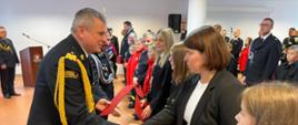 Pomorski komendant wojewódzki Państwowej Straży Pożarnej uściska dłoń kobiecie w drugiej trzyma czerwoną teczkę obok stoją inni przedstawiciele Młodzieżowych Drużyn Pożarniczych.