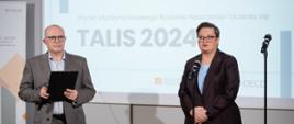Dwóch prelegentów na scenie podczas konferencji dotyczącej wyników badania TALIS 2024. Mężczyzna w szarym garniturze trzyma czarną teczkę po lewej, a kobieta w ciemnym garniturze i okularach stoi przy mikrofonie po prawej. W tle widoczny jest ekran z tytułem: Nauczycielki i nauczyciele o pracy w zawodzie, Wyniki Międzynarodowego Badania Nauczania i Uczenia się oraz duży napis TALIS 2024."