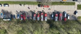Widok z lotu ptaka na zgrupowanie służb ratowniczych. Wzdłuż drogi zaparkowanych jest kilkanaście pojazdów: czerwone wozy strażackie, białe karetki pogotowia oraz radiowozy policji. Przy pojazdach widać grupy ratowników.