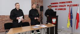dwóch funkcjonariuszy ubranych w ubrania koszarowe państwowej straży pożarnej stoi przed stołem na którym leżą otwarte dokumenty jeden strażak stoi po prawej stronie przed trybuną na której stoi laptop osoba z lewej strony trzyma w dłoniach białą kartkę a osoba po jej lewej stronie długopis w tle ekran projekcyjny na tle z jasnoczerwonej cegły w prawym narożniku stoi kolumna na statywie na prawej ścianie widoczny duży czerwony napis komenda powiatowa państwowej straży pożarnej w Łowiczu a przed napisem na podłodze stoi stojak a w nim trzy flagi flaga polski miasta Łowicz oraz unii europejskiej na podłodze panele barwy jasnoszarej