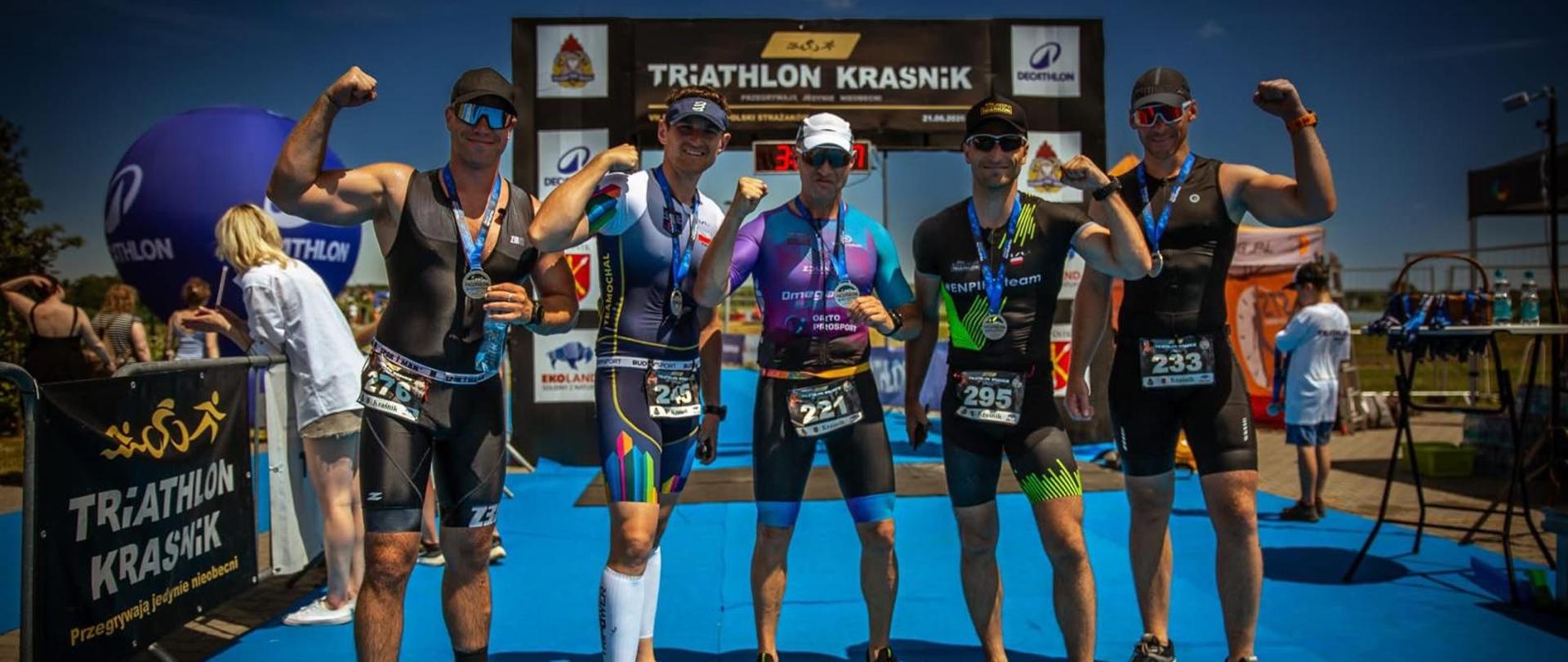 Pięć osób w strojach startowych frontem do obiektywu, w tle meta zawodów triathlonowych