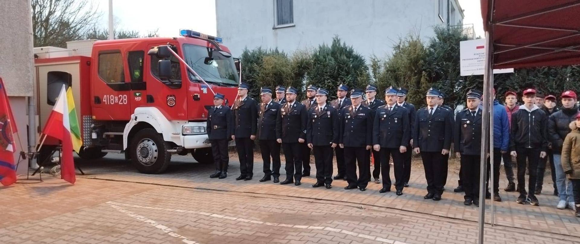 Widok na pododdział druhów OSP - ok. 20 osób. Obok nie w pełni widoczny pojazd pożarniczy, udekorowany uroczyście. Z lewej strony fragment budynku i stojak z flagami - narodowa, Związku ZOSP RP, gminna.