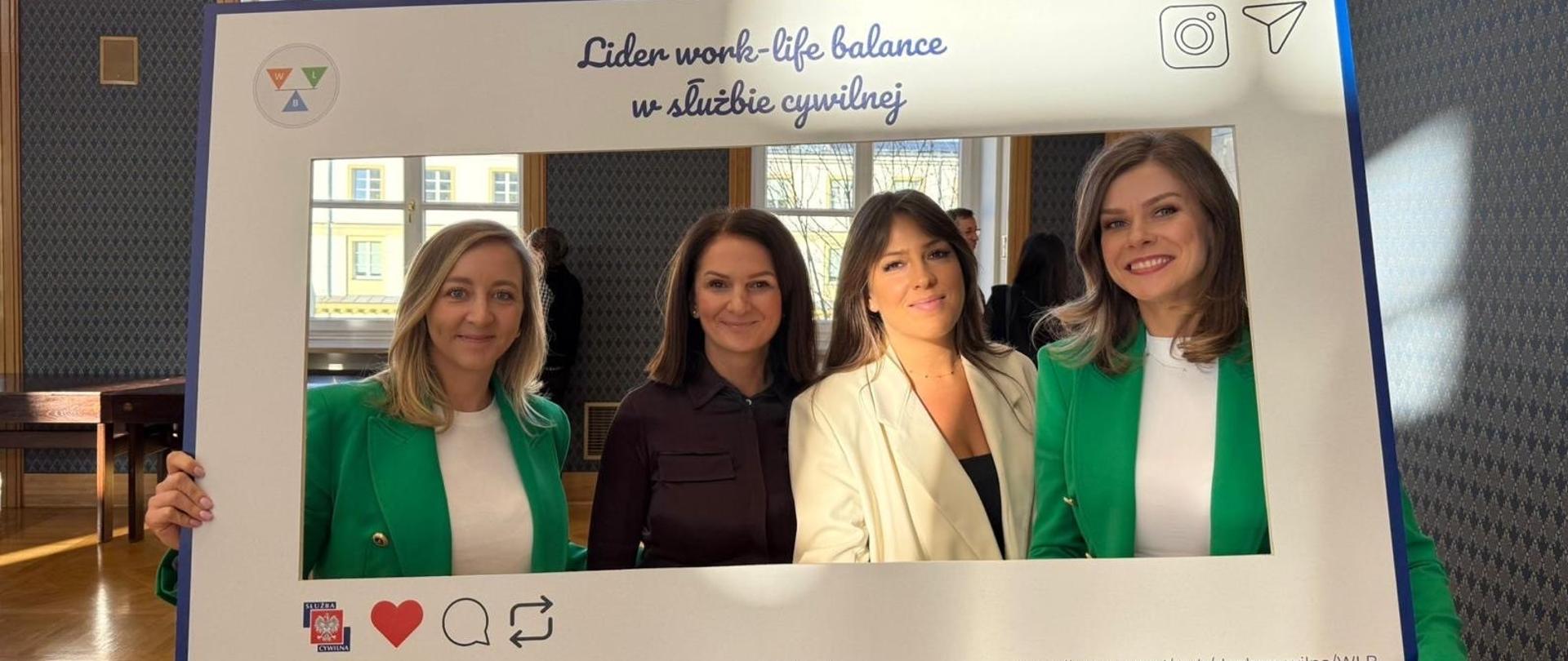 Na zdjęciu widać cztery kobiety pozujące za dużą ramką. Kobiety stoją obok siebie "wewnątrz ramki". Wszystkie są uśmiechnięte. Dwie skrajne kobiety (pierwsza od lewej i pierwsza od prawej) są ubrane w zielone marynarki. Druga kobieta od lewej ma na sobie ciemną koszulę, a trzecia od lewej jest ubrana w jasny, kremowy żakiet. Ramka jest duża, biała imitująca post z mediów społecznościowych. Na ramce znajdują się napisy: "#służbacywilna #WorkLifeBalance #RównowagaToSiła #Wellbeing. 