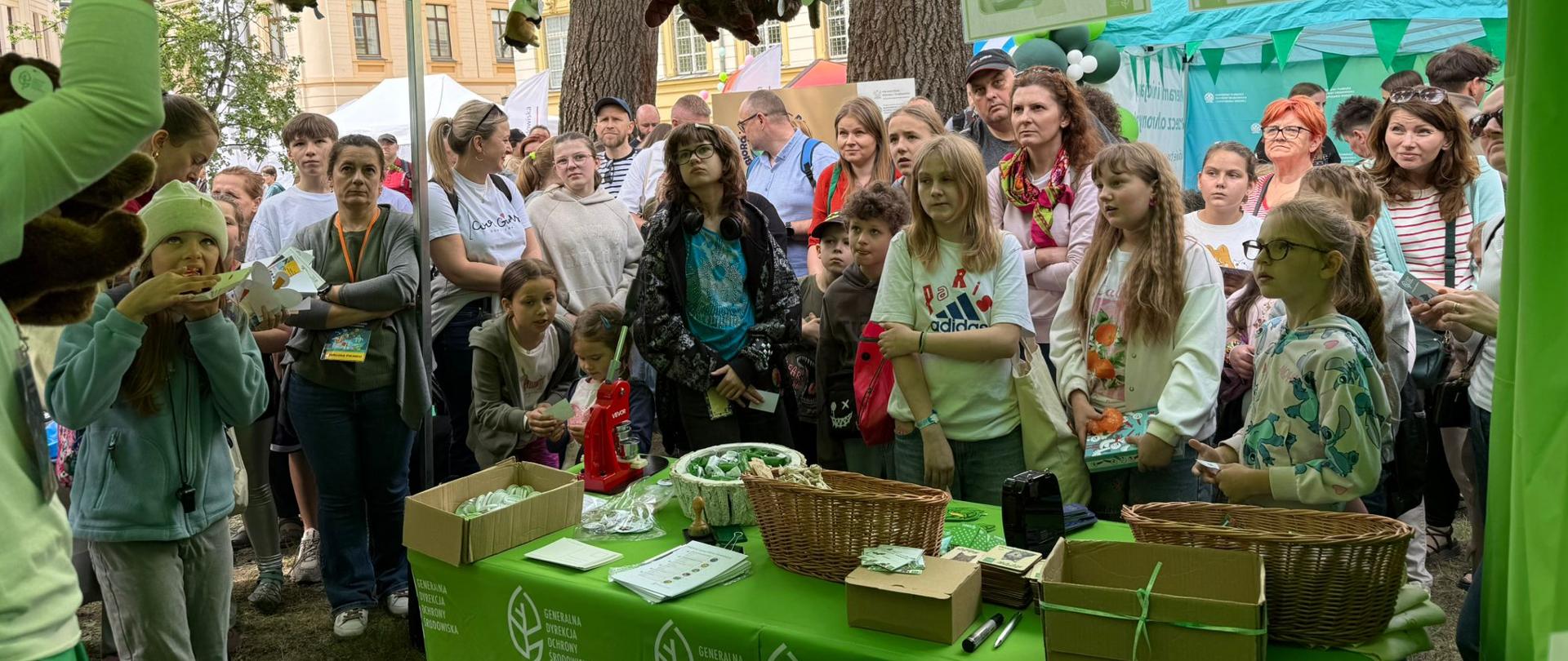 Zielone stoisko Generalnej Dyrekcji Ochrony Środowiska (namiot ozdobiony logotypami - białe liście). Na stole wyłożone materiały promocyjne. Dookoła stołu stoi grupa dorosłych z dziećmi.