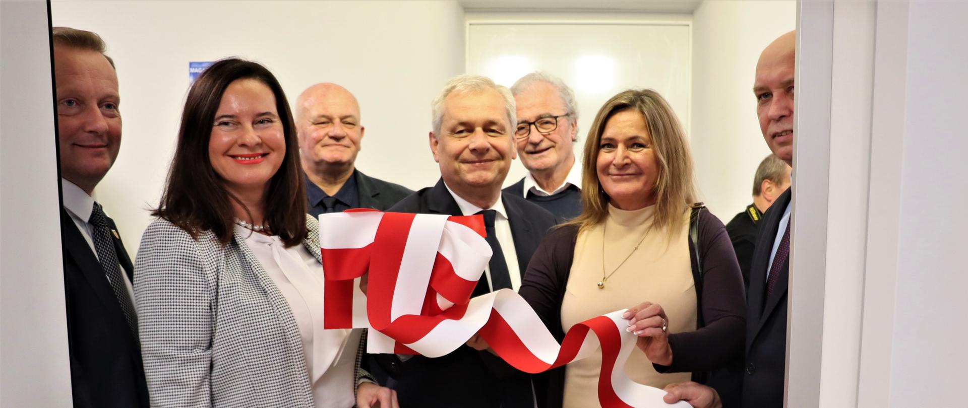 Dwie kobiety oraz piątka mężczyzn stojących w białym holu ze wstęgą