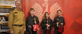 Strażak w mundurze stoi obok trójki dzieci w strojach strażackich, które trzymają puchary i czerwone torby z nagrodami. Zdjęcie wykonano na tle czerwonej ścianki z symbolem straży pożarnej i informacjami o jednostce. Dzieci wyglądają na dumne i zadowolone, a cała scena ma charakter uroczystego wręczenia nagród.