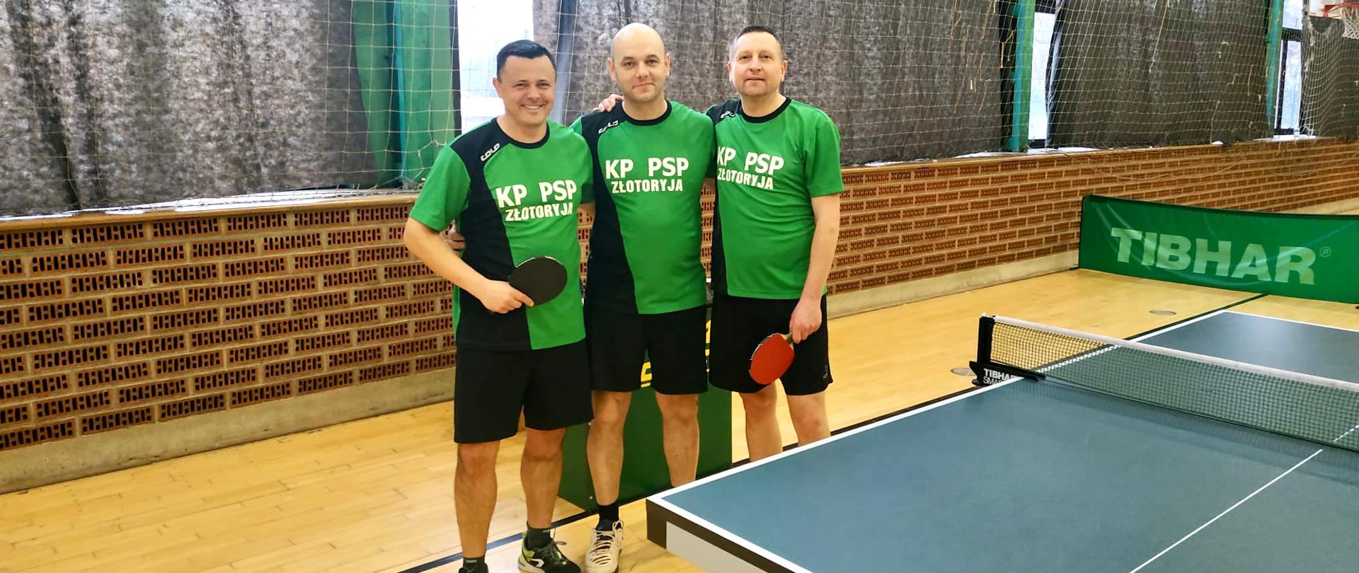 trzech młodych mężczyzn ubranych w zielono- czarne stroje sportowe w obiekcie sali gimnastycznej, po prawej stronie widoczny stół do tenisa stołowego