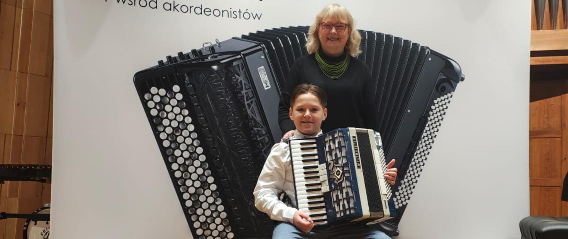 Kolorowe zdjęcie. Na scenie na tle białego baneru z czarnym akordeonem siedzi na krześle Bartosz i trzyma akordeon a za nim stoi Pani Adriana Brola