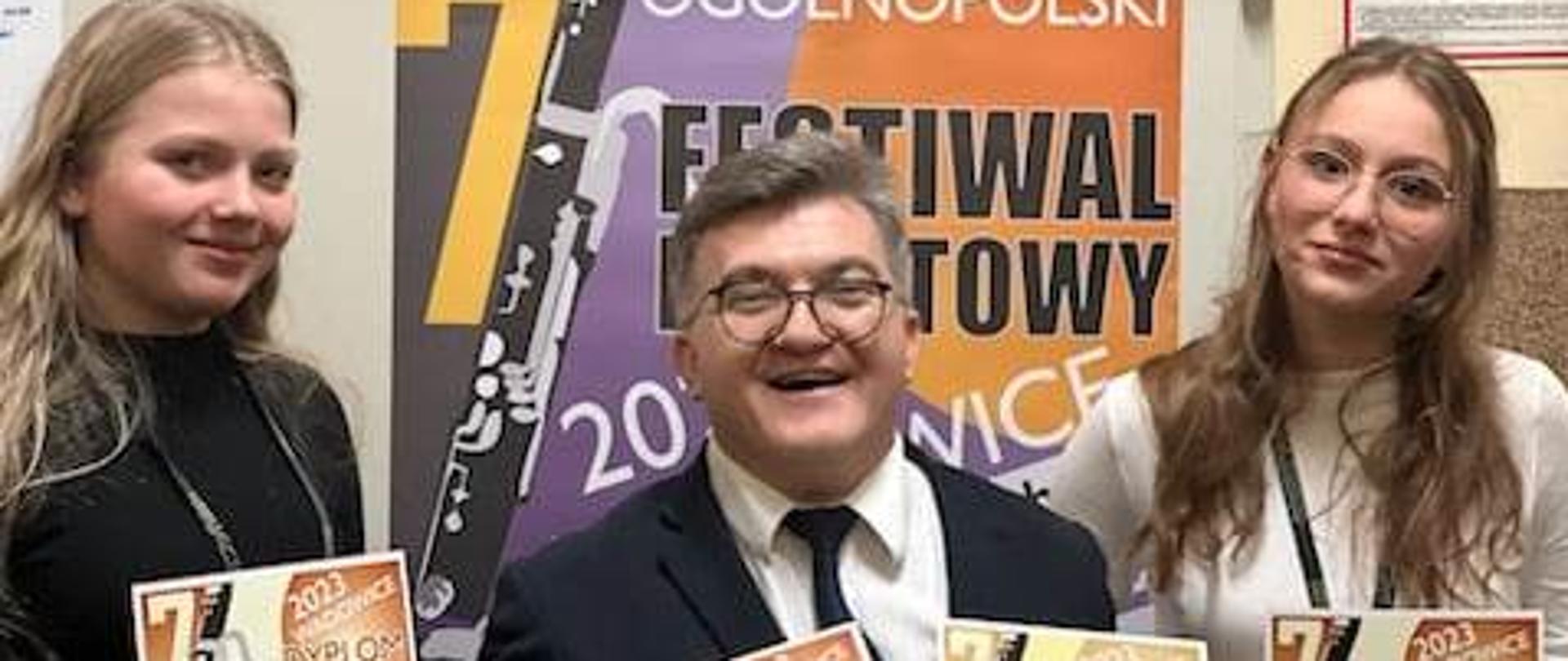 zdjęcie przedstawia uczennice naszej szkoły z dyplomami z 7 Ogólnopolskiego Festiwalu Fagotowym w Wadowicach, po lewej i po prawej stronie zdjęcia oraz ich nauczyciela Marcina Pokusę w środku, także z dyplomami w ręku, w tle widać baner promujący konkurs oraz beżową ścianę