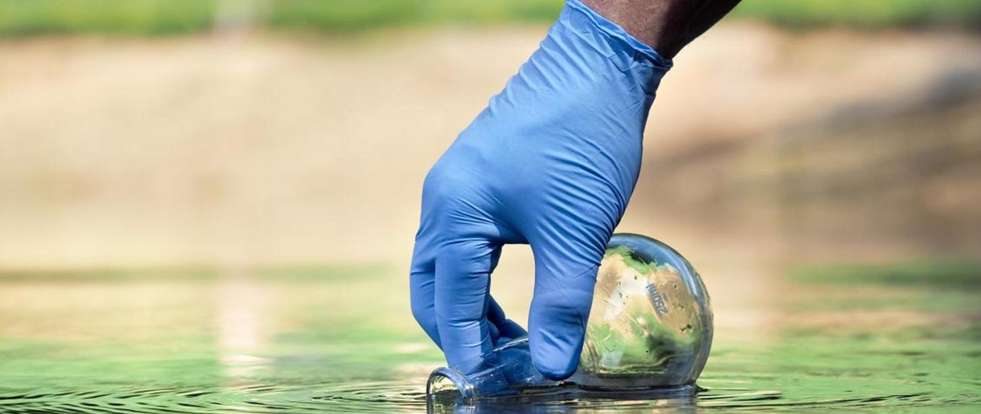 Dłoń w niebieskiej rękawiczce ochronnej nabiera wodę z miejsca poboru do szklanego naczynia