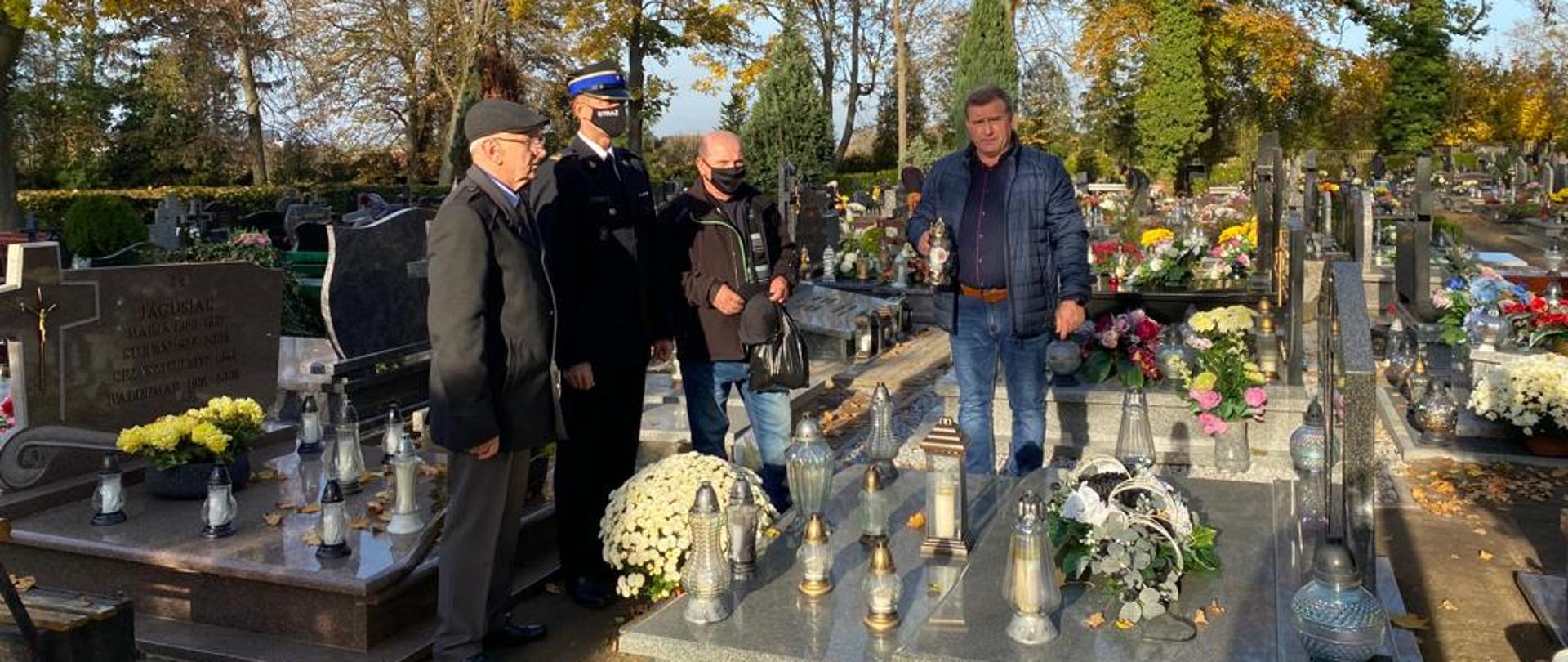 Na zdjęciu widzimy 4 mężczyzn stojących przy grobie na cmentarzu. Jeden z nich ubrany w mundur strażacki a pozostali w ubraniach cywilnych.