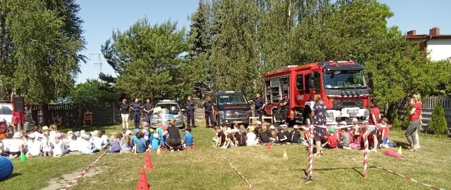 Strażacy oraz dzieci przedszkolne z opiekunami uczestniczą w zajęciach związanym z bezpieczeństwem.