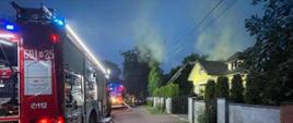 Samochód pożarniczy stoi na ulicy przy chodniku. Widać rozwinięte węże z wodą i dom z prawej strony. Nad domem unosi się dym.