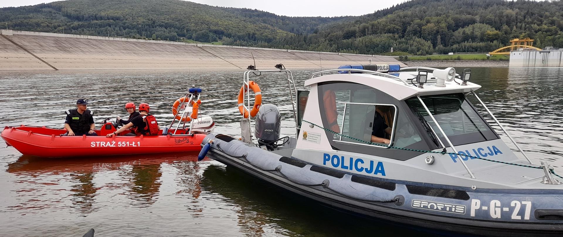 Widok z przodu. Po lewej stronie w łodzi motorowej PSP siedzi policjant i dwóch strażaków. Po prawej stronie widoczna łódź motorowa Policji. W tle widoczna zapora wodna.