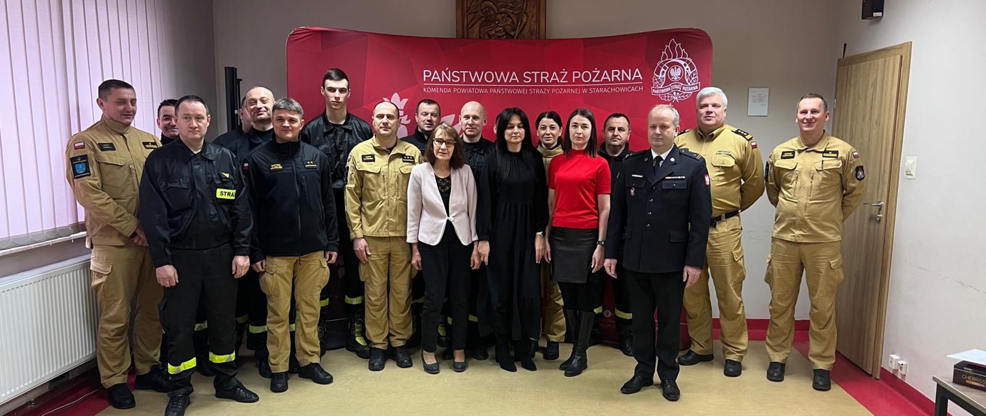 Strażacy na świetlicy, strażacy pozują do zdjęcia na czerwonym tle z logiem psp. 