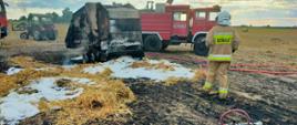 Na zdjęciu widać spaloną prasę rolniczą. W tle widać samochody strażackie oraz ciągnik. Przed prasą stoi strażak z wężem. Za nim przygotowane jest urządzenie do podania piany gaśniczej