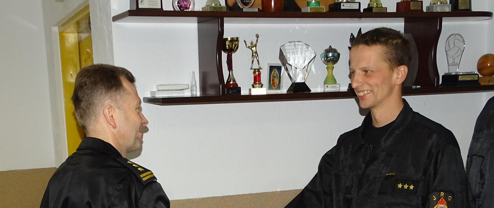 Zdjęcie wykonano wewnątrz budynku komendy powiatowej Państwowej Straży Pożarnej w pomieszczeniu świetlicy. Komendant Powiatowy Państwowej Straży Pożarnej w sępólnie krajeńskim gratuluje wyróżnionemu strażakowi ściskając mu dłoń. 