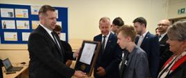 Minister Czarnek podaje młodemu chłopakowi w szarek koszuli dyplom oprawiony w ramki, obok stoi grupa ludzi.