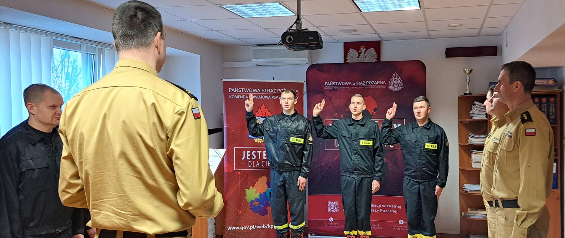 Trzech funkcjonariuszy z podniesioną prawą ręką składa ślubowanie przed Komendantem Powiatowym w sali Komendy. 