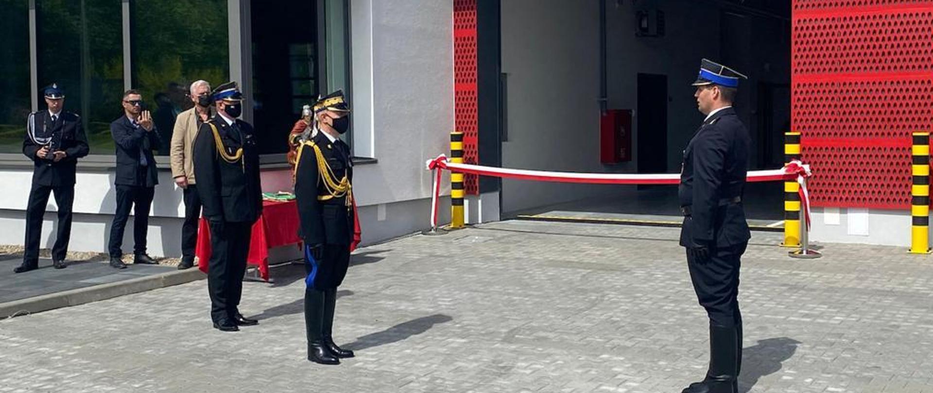 po prawo stoi na baczność funkcjonariusz PSP po przeciwnej stronie stoi zwrócony w jego stronę komendant główny PSP, za nim kilku mężczyzn stoi, w dalszym tle widać biało - czerwoną wstęgę, która będzie przecięta symbolicznie na otwarcie nowo wybudowanej JRG 1 w Koszalinie