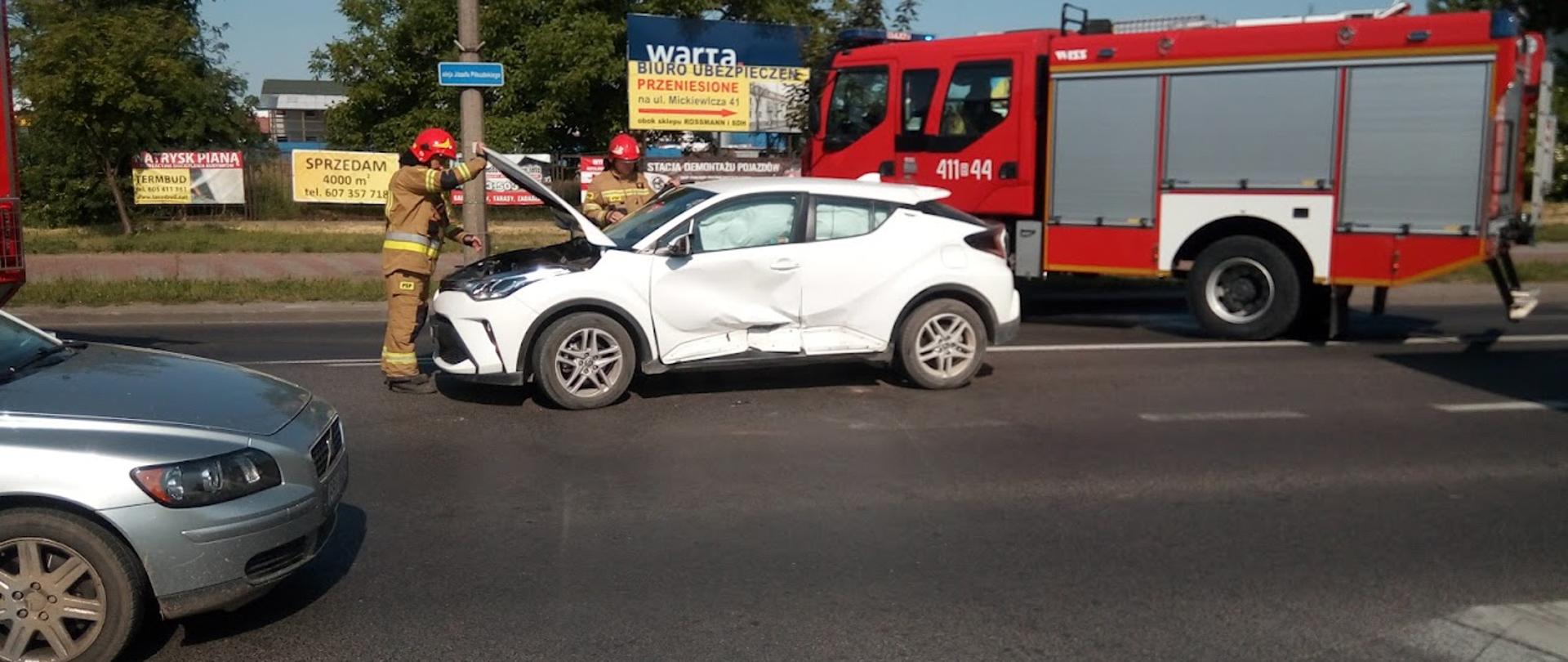 Zdjęcie przedstawia dwóch strażaków przy białym samochodzie osobowym po kolizji z innym autem z podniesioną pokrywą maski na środku jezdni, obok stoi czerwony pojazd ratowniczo-gaśniczy