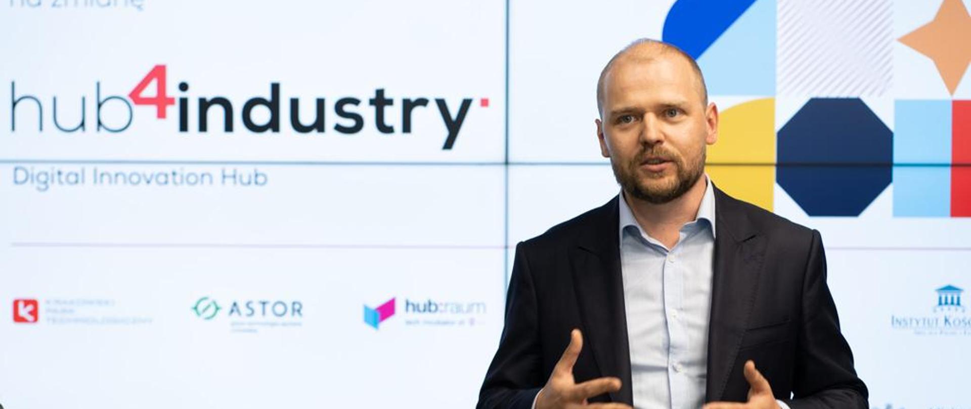 Minister Krzysztof Mazur podczas przemówienia; gestykuluje rękami; za jego plecami ekran z kolorową grafiką i napisem Hub4Industry.