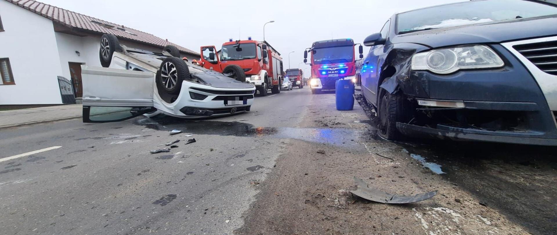 Biały samochód leży na dachu z otwartymi drzwiami za samochodem białym stoją trzy samochody strażackie oraz jeden policyjny w górnym prawym rogu widoczny granatowy samochód z rozbitym przodem na drodze wycieki płynów eksploatacyjnych