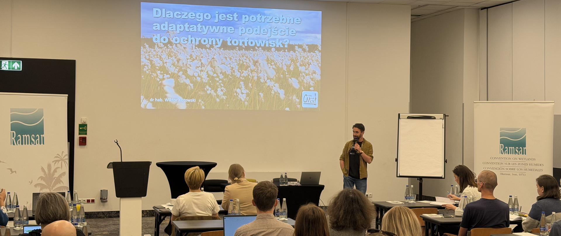 Sala konferencyjna. Przy stolikach siedzą uczestnicy. Na środku sali stoi mężczyzna z mikrofonem. Za jego plecami znajduje się ekran. 
