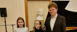Zdjęcie przedstawiające wykonawców Zofię Tambur i Julię Buchowską wraz ze swoim nauczycielem panem Mikołajem Potoczakiem, którzy wystąpili w Zespole Szkół Muzycznych im. Fryderyka Chopina w Legnicy. Dziewczęta zagrały w sali koncertowej podczas koncertu zorganizowanego w ramach pianistycznych prezentacji talentów klas pierwszych.