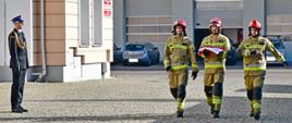 Trzech funkcjonariusze Państwowej Straży Pożarnej w umundurowaniu specjalnym oraz hełmie maszerują, środkowy strażak niesie flagę. Niedaleko stoi funkcjonariusz Państwowej Straży Pożarnej w umundurowaniu wyjściowym.