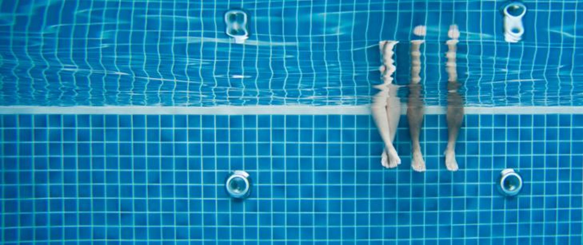 Zdjęcie zrobione na basenie pod wodą. Widać na nim dwie pary nóg.