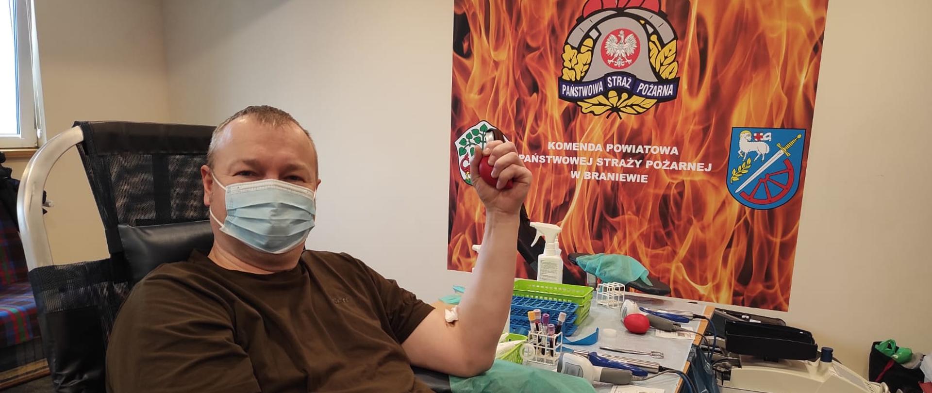 Na zdjęciu pan w maseczce. Siedzi na fotelu w trakcie pobierania krwi. W tle logo PSP i płomienie