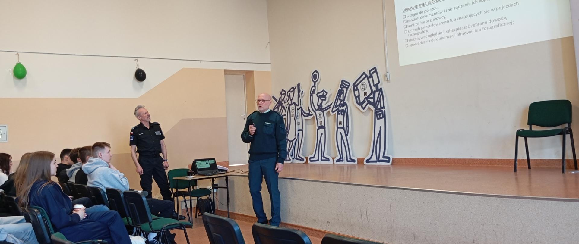 Sala wykładowa w szkole. Inspektor prowadzący prelekcję oraz kilku uczniów siedzących na widowni słuchających wykładu
