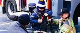 Na zdjęciu widzimy chłopca ubierającego aparat ODO na plecy. Pomaga mu w tym strażak oraz opiekun.