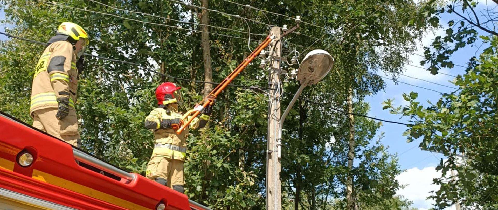Strażacy w ubraniach specjalnych oraz hełmach z opuszczonymi przyłbicami stoją na dachu samochodu pożarniczego. Jeden z nich przy pomocy izolowanego drążka dielektrycznego zakończonego nożycami do cięcia przewodów odcina przewód elektryczny linii niskiego napięcia. 
