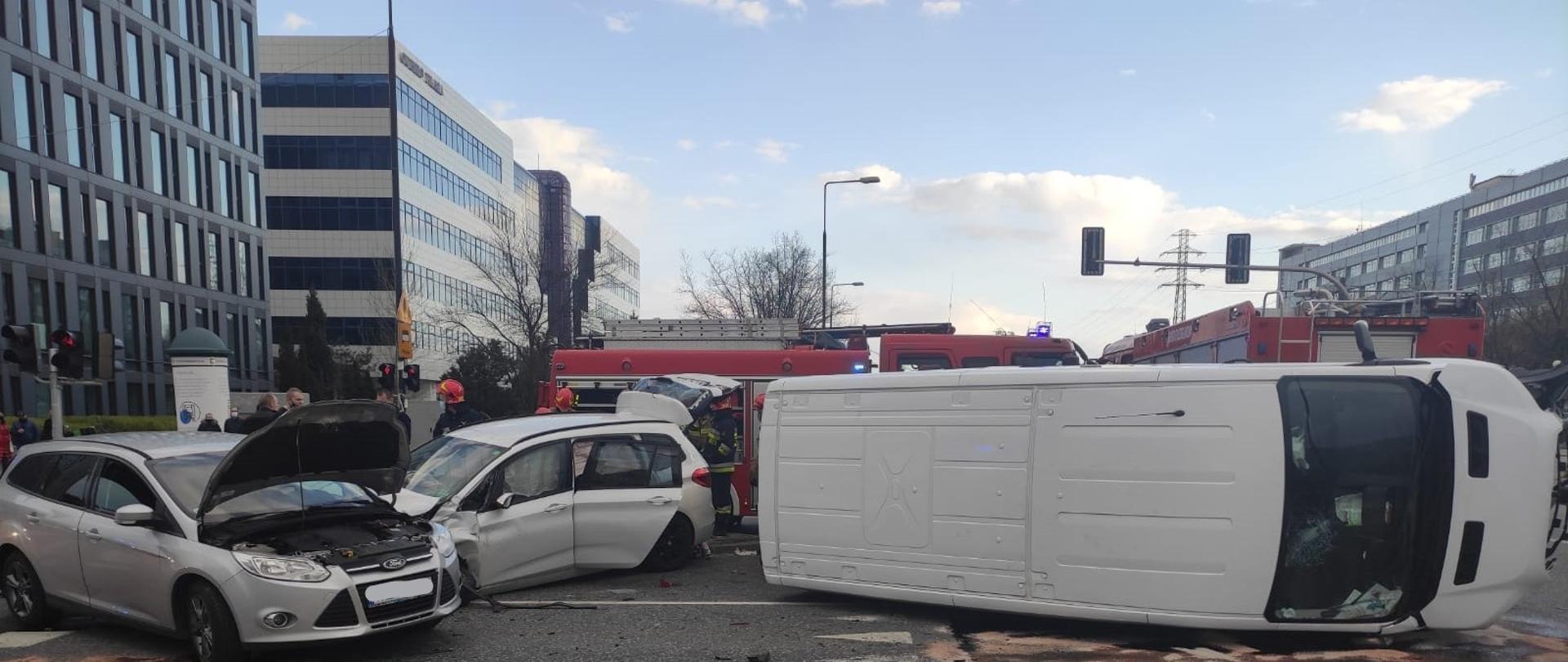 Zdjęcie 1 – Na zdjęciu znajdują się trzy uszkodzone samochody biorące udział w wypadku, jeden z pojazdów ma otwartą pokrywę silnika, samochód odstawczy leży na prawym boku. W tle widoczne są budynki biurowe