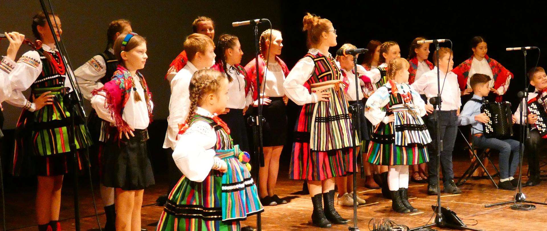Zdjęcie przedstawiające stojących na scenie kilkunastu uczniów szkoły muzycznej w Opocznie. Są oni ubrani w stroje ludowe.