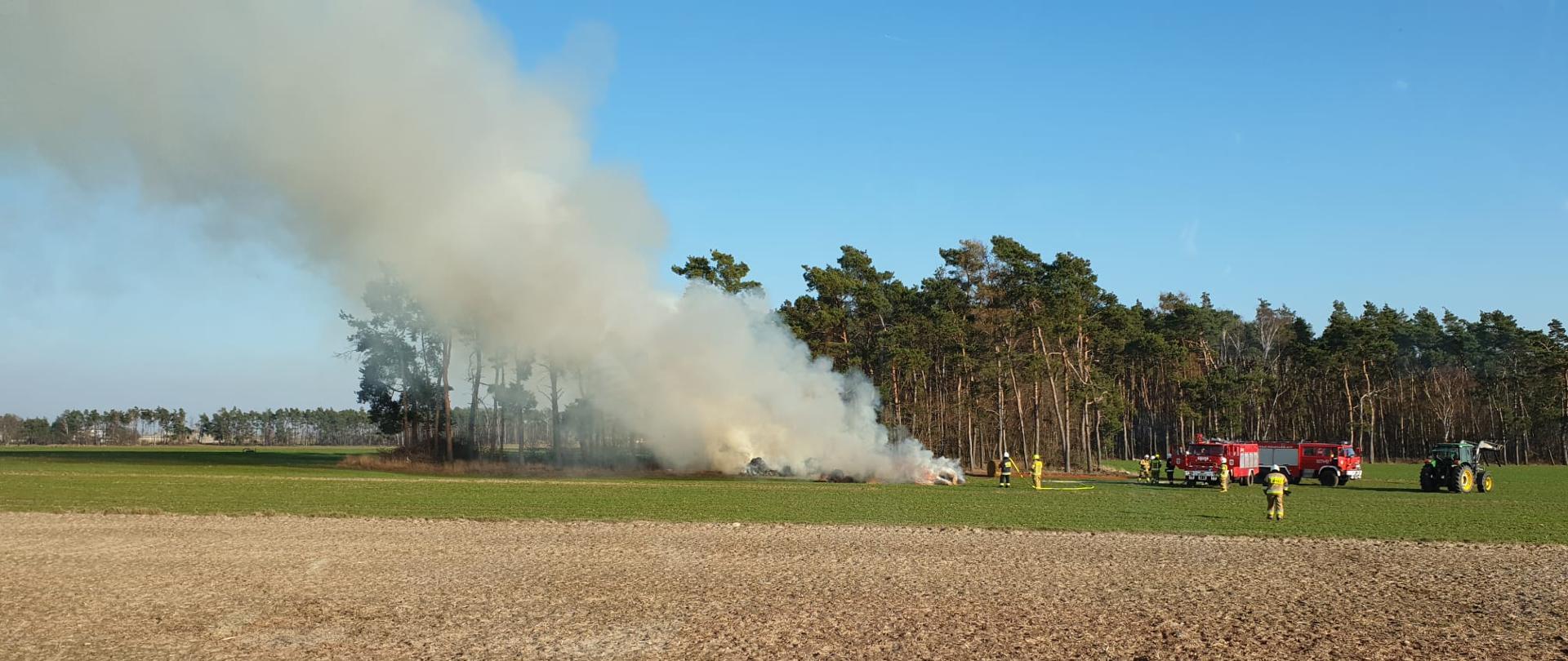 Na zdjęciu palące się baloty słomy na polu w oddali samochody gaśnicze oraz ciągnik i ratownicy podczas działań