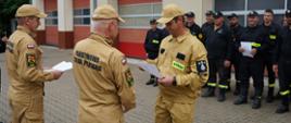 Na placu przed komendą, na tle garaży stoją uczestnicy szkolenia. Na przeciwko dwóch strażaków PSP w mundurach służbowych koloru piaskowego. Stojący z prawej strony zastępca komendanta wręcza zaświadczenie jednemu ze słuchaczy.