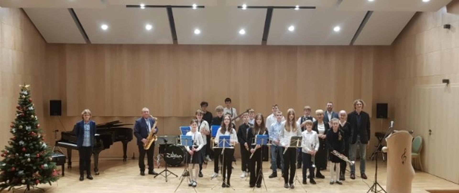Zdjęcie przedstawia scenę, na której stoi zespół muzyczny składający się z nauczycieli i uczniów szkoły. Każdy trzyma w ręku instrument: saksofon, flet, trąbkę, gitarę. Po lewej stronie w rogu sceny stoi udekorowana choinka a za nia czarny fortepian.