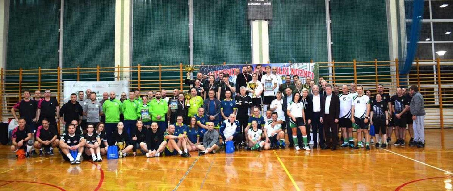Zdjęcie przedstawia dużą grupę osób, uczestników V Turnieju Halowej Piłki Nożnej Kadr Szkół Rolniczych o Puchar Ministra Rolnictwa i Rozwoju Wsi, pozujących do wspólnego zdjęcia w hali sportowej Zespołu Szkół Centrum Kształcenia Rolniczego w Siennicy Różanej.
Grupa i kontekst: Na zdjęciu jest kilkadziesiąt osób, w tym kadra nauczycielska, trenerzy i zawodnicy z różnych szkół rolniczych z całej Polski. Uczestnicy stoją w kilku rzędach na drewnianym parkiecie, przed bramką do piłki ręcznej z biało-czerwonymi słupkami. Wszyscy wydają się zadowoleni.
Ubiór: Wiele osób ubranych jest w stroje sportowe reprezentujące różne drużyny (np. zielone bluzy, czarno-białe stroje). Część osób ma zawieszone na szyi medale na czerwonych wstążkach.
Nagrody i tło: Na środku, na podłodze, widoczne są puchary i nagrody. Bezpośrednio za grupą zawieszony jest duży baner z nazwą i logiem turnieju oraz szkoły gospodarza (m.in. ""URNIEJ"", ""MINISTRA ROLNICTWA"", ""Siennica Różana""). W tle widoczna jest zielono-żółta ściana hali sportowej i siatki zabezpieczające.