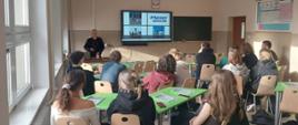 na zdjęciu młodzież siedzi przy stolikach lekcyjnych, przed stolikami na ścianie wisi ekran multimedialny na którym jest wyświetlana prezentacja którą omawia pracownica Powiatowej Stacji Sanitarno-Epidemiologicznej w Szczytnie