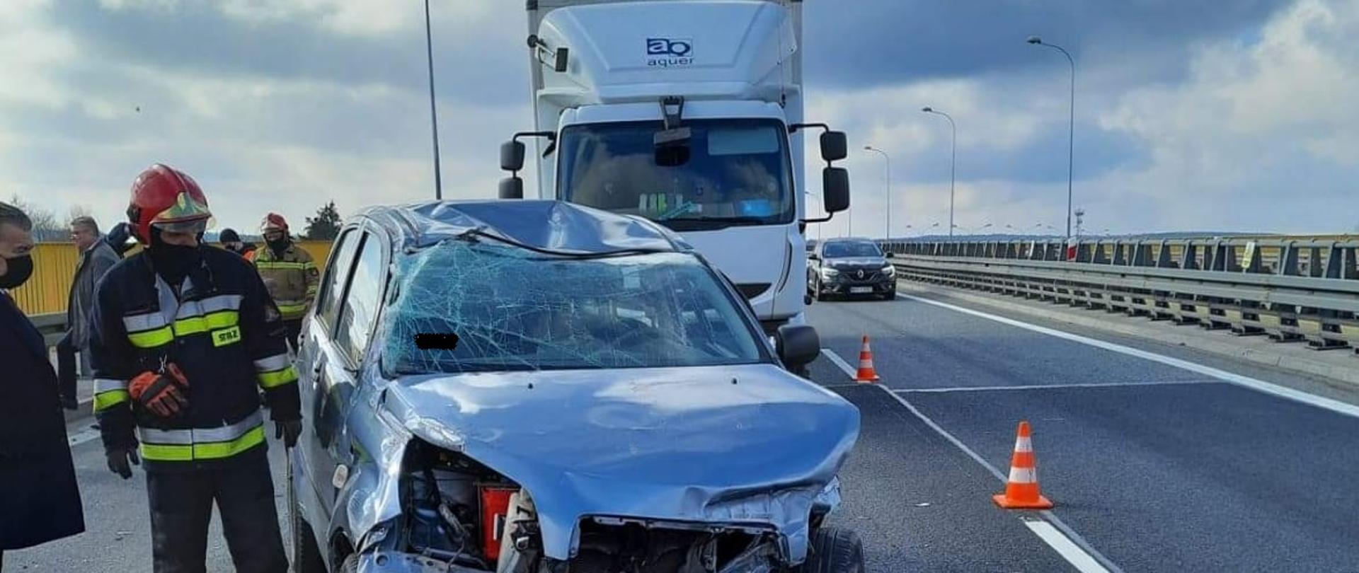 Na zdjęciu widzimy uszkodzony przód samochodu osobowego DAIHATSU z którego wyciekły płyny eksploatacyjne na pas jezdni. Samochód w wielu miejscach ma pogniecioną karoserię. Szyba z przodu jej popękana. Za nim widać samochód ciężarowy RENAULT