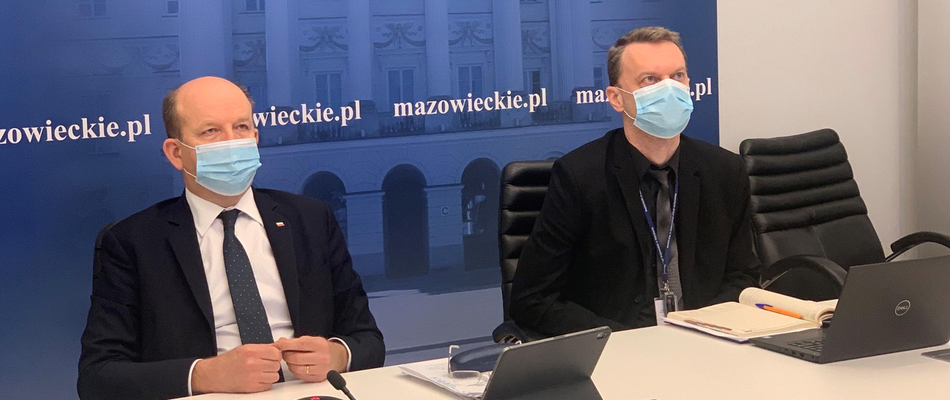 Na zdjęciu znajduje się dwóch elegancko ubranych mężczyzn, którzy siedzą za stołem i biorą udział w wideokonferencji