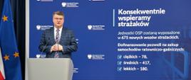 Maciej Wąsik przemawia przy mównicy w trakcie konferencji prasowej za nim banner mswia i napisami konsekwentnie wspieramy strażaków. Jednostki OSP zostaną wyposażone w 675 nowych wozów strażackich. dofinansowanie pozwoli na zakup samochodów ratowniczo-gaśniczych ciężkich 78 średnich 417 lekkich 180
