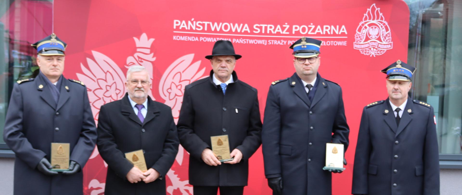 strażacy stoją przed czerwonym banerem z orłem trzymając w ręku paiątkowe statuetki
