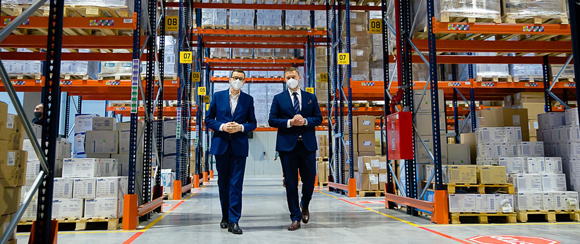 Premier Mateusz Morawiecki z prezesem firmy idą pomiędzy regałami.