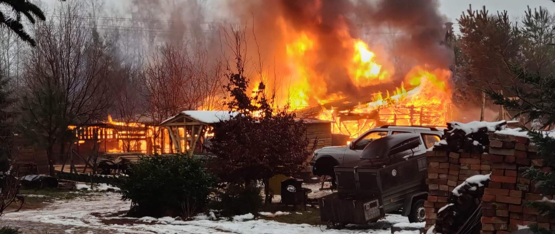 Dzień. Na ziemi leży śnieg. Widok na część podwórza/ogrodu. Na pierwszym planie kilka niskich drzewek, z prawej strony ułożony stos ze starych cegłówek, obok którego stoi przyczepka samochodowa. Za przyczepką widać samochód osobowy, kombi w kolorze białym. W tle za pojazdem i drzewkami widać całkowicie objęte pożarem drewniany obiekt. Ogień wznosi się wysoko na wiatą. Widać dużo czarnego dymu