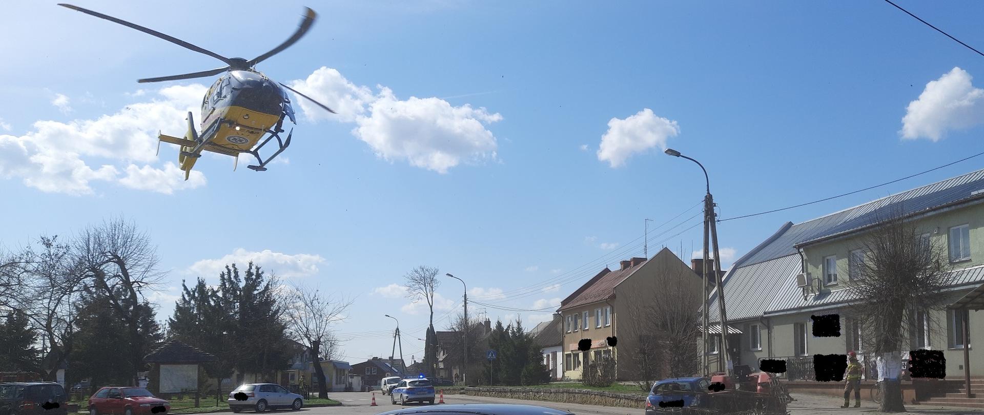 Zdjęcie obrazuje kilka samochodów osobowych ciągnik rolniczy lądujący śmigłowiec LPR, radiowóz, strażaka, budynki i drzewa w tle