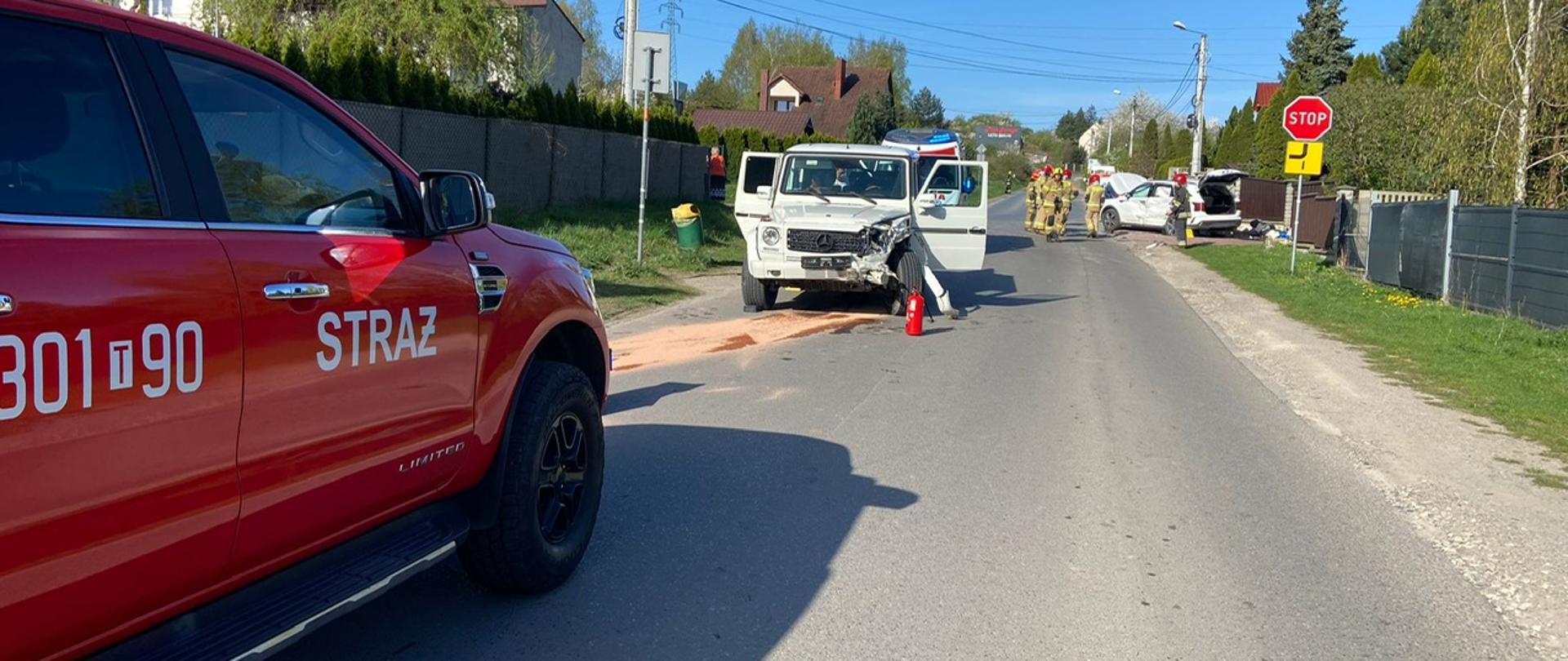 Zdjęcie przedstawia ulicę Prostą gdzie doszło do zderzenia pojazdów. Po lewej stronie stoi samochód operacyjny Państwowej Straży Pożarnej, dalej pierwszy z uszkodzonych aut – terenowy mercedes, za którego wystaje ambulans Państwowego Ratownictwa Medycznego. Po drugiej stronie ulicy, na poboczu stoi drugie zniszczone w wyniku zderzenia auto – Kia Sorento. Na jezdni stoją strażacy.